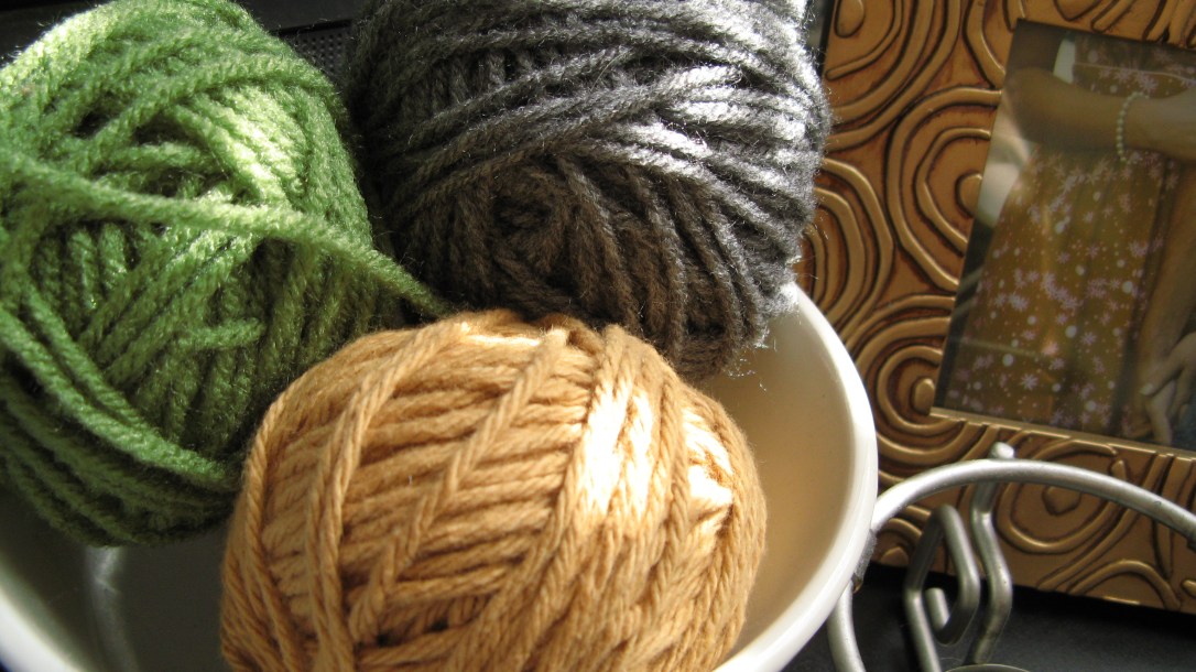 white ceramic bowl holding three balls of yarn - green, light brown, and grey - next to a gold picture frame white ceramic bowl holding three balls of yarn - green, light brown, and grey - next to a gold picture frame