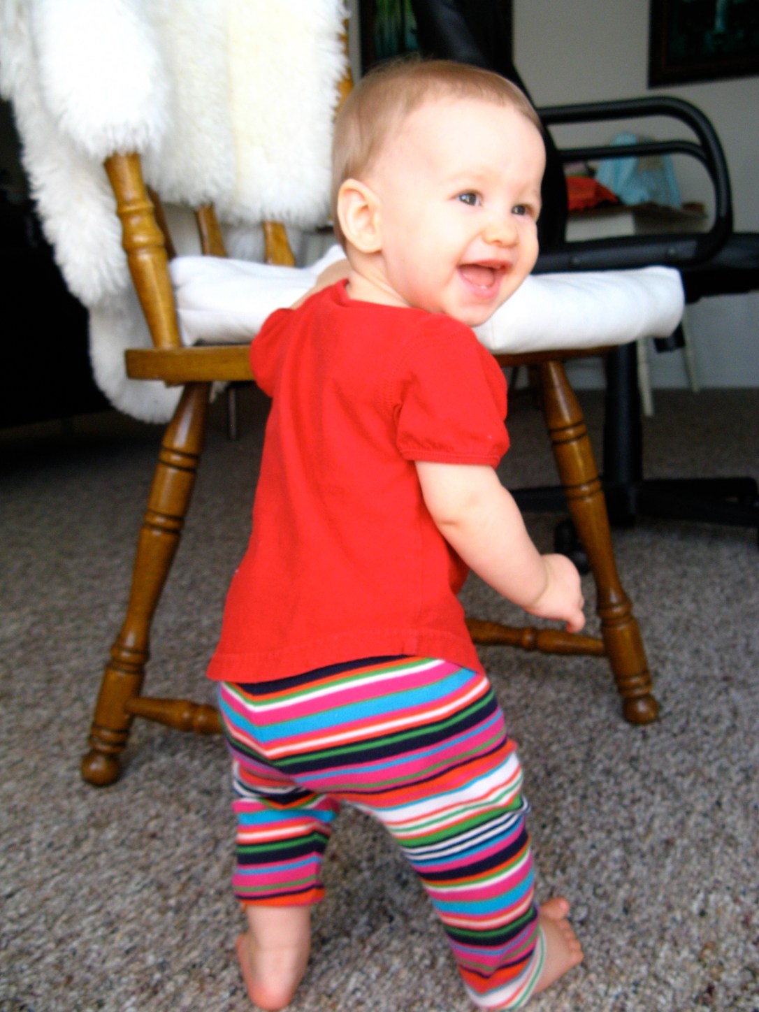 Happy baby wearing striped leggings standing up next to wooden chair with IKEA Rens sheepskin and chair cushion