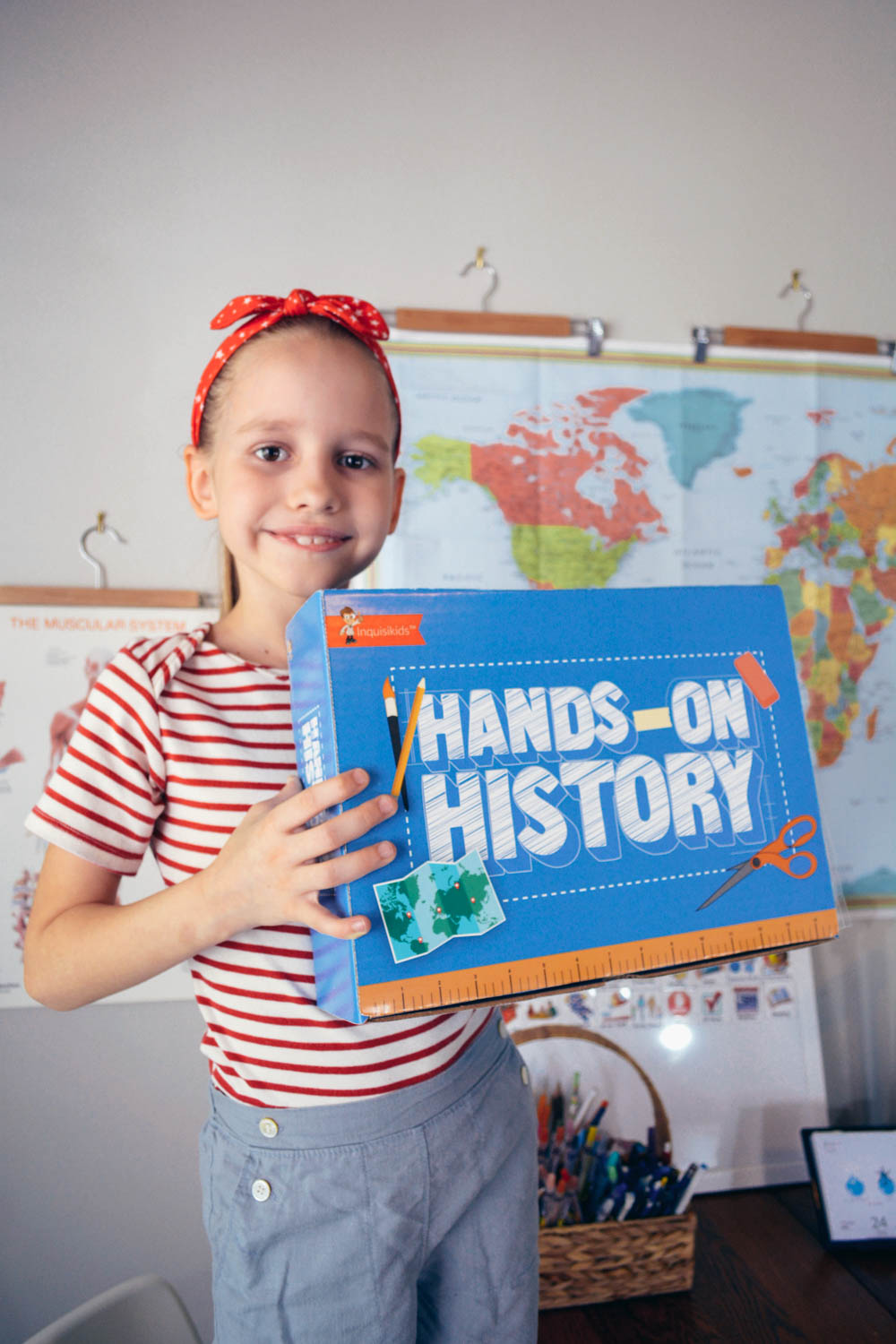 A student stands in front of a world map, holding a Sonlight Hands-on History box 
