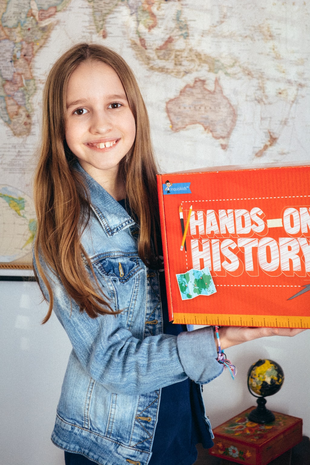 A student stands in front of a world map, holding a Hands-on History box from Sonlight 