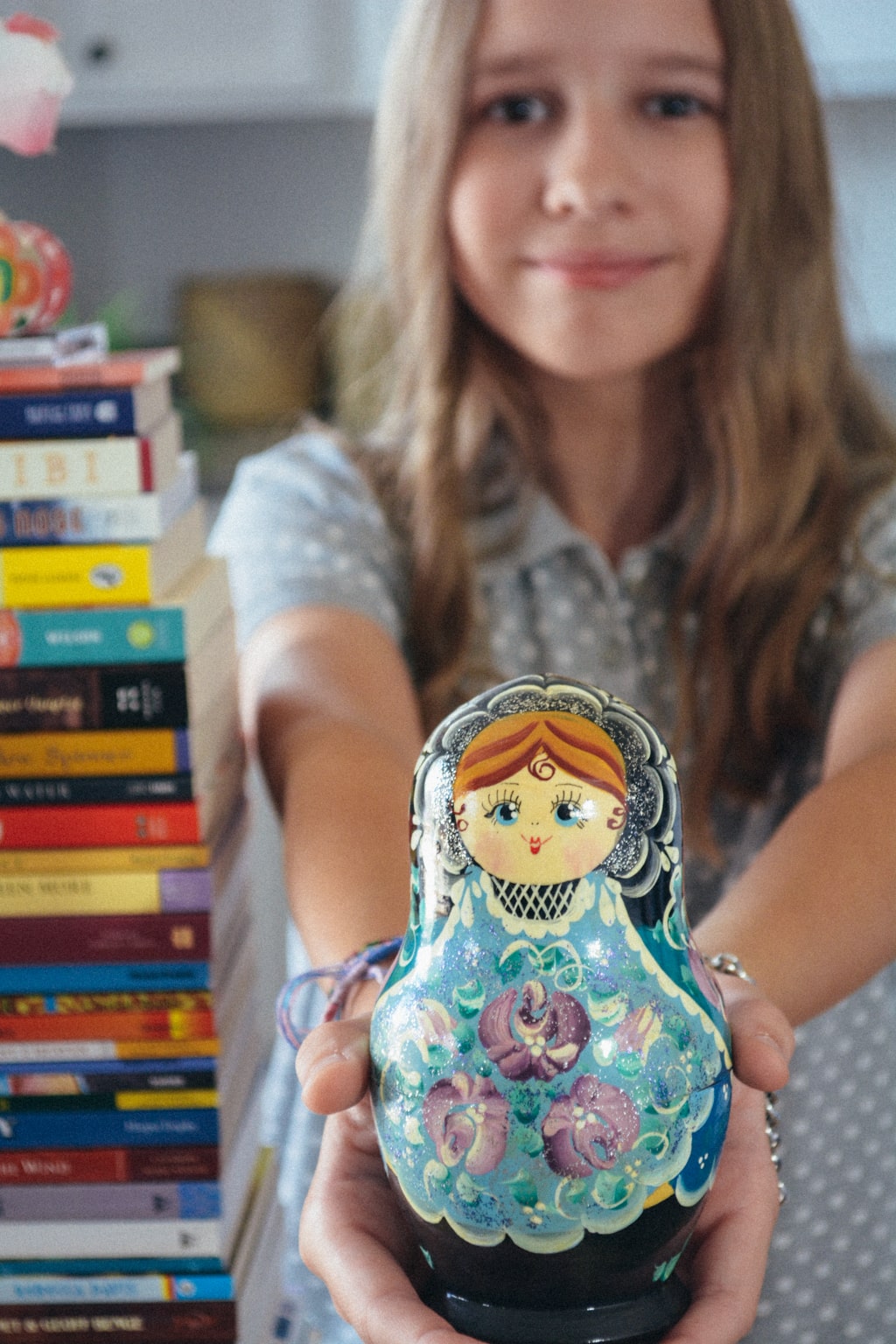 Homeschool student stands next to a very tall stack of diverse middle grade books, while holding a hand-painted matryoshka toward the camera